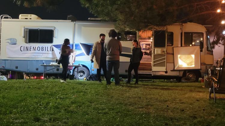 Night scene. RV with a “Cinemobilia” sign on the side. 4 people staging in front talking, while another is working on a fold-up table.